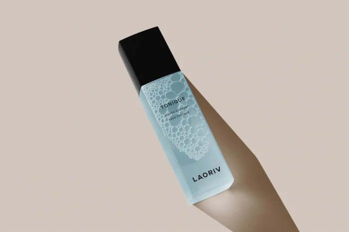 Minimalist cosmetic bottle mockup featuring a translucent blue toner bottle with bubbles inside, black cap, and shadow cast on a beige background